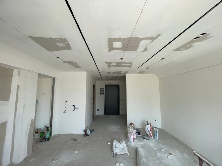 Empty unfinished room with drywall, exposed ceiling fixtures, and construction materials scattered on the floor