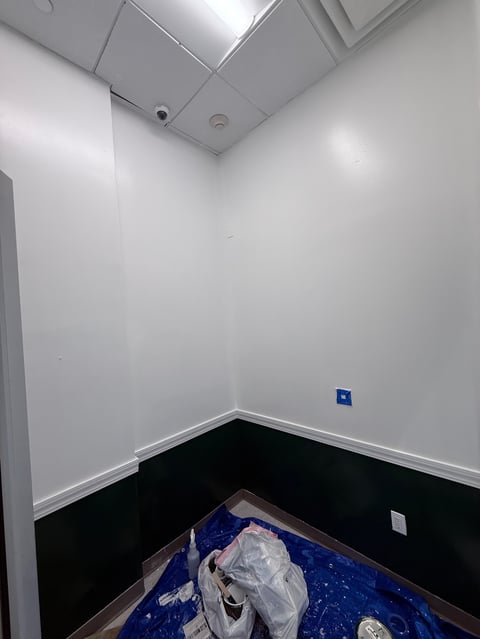 Empty room with white walls, drop ceiling, black baseboard, and blue protective floor covering with construction materials