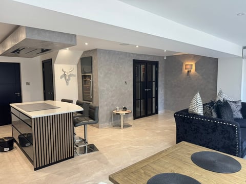 Modern open-plan apartment interior with kitchen island, dark sofa, light wood flooring, and contemporary furnishings