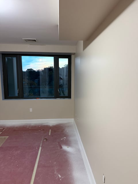 Empty room under construction with beige walls, protective flooring, and large black-framed window overlooking a city skyline at dusk