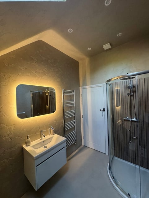 Modern bathroom with illuminated mirror above white vanity sink, heated towel rack, and corner shower enclosure with dark glass doors