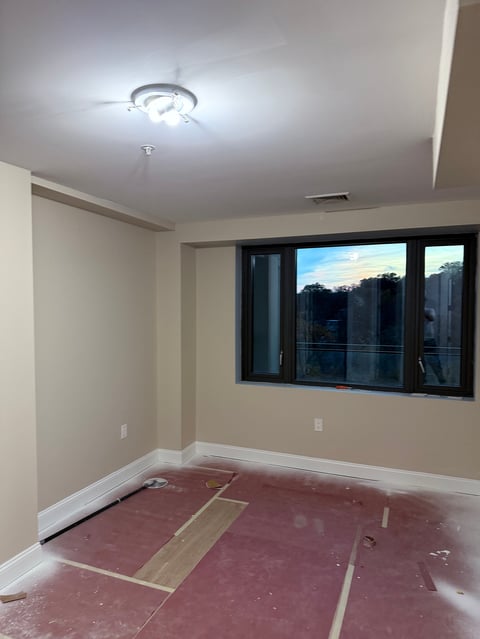 Empty room under renovation with beige walls, red subfloor, ceiling light fixture, and large windows overlooking trees at dusk